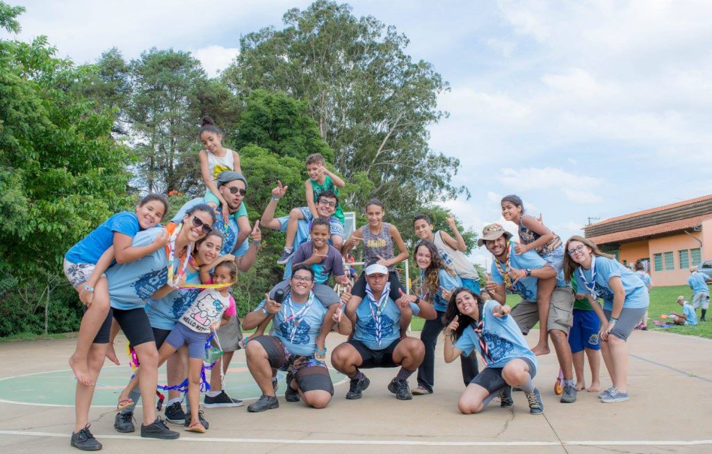 Foto em ambiente aberto com árvores ao fundo e céu claro com algumas nuvens. À frente há um grupo de jovens e chefes em pose para a foto, usam lenços diferentes, camiseta azul clara, bermuda e tênis, há algumas crianças que não usam lenço e nem a camiseta azul.