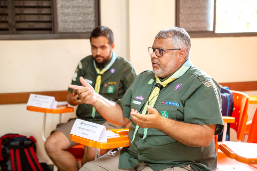 Ao fundo da foto uma parede bege com duas janelas. Algumas cadeiras universitárias e laranjas, em duas, dois chefes sentados. Ambos usam o vestuário escoteiro e lenço da nacional e possuem um papel dobrado em triângulo na frente de cada um. O chefe em destaque é pardo, tem cabelo e barba branca, usa óculos de grau, a boca está um pouco aberta, seus braços estão dobrados a 90 graus e as mãos estão abertas e voltadas com a palma para cima.